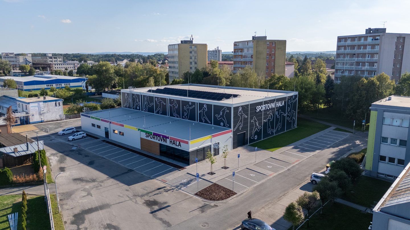 Pohled z výšky od sídliště: hala mezi panelovými domy a zelení, s barevným pásem nad vstupem.