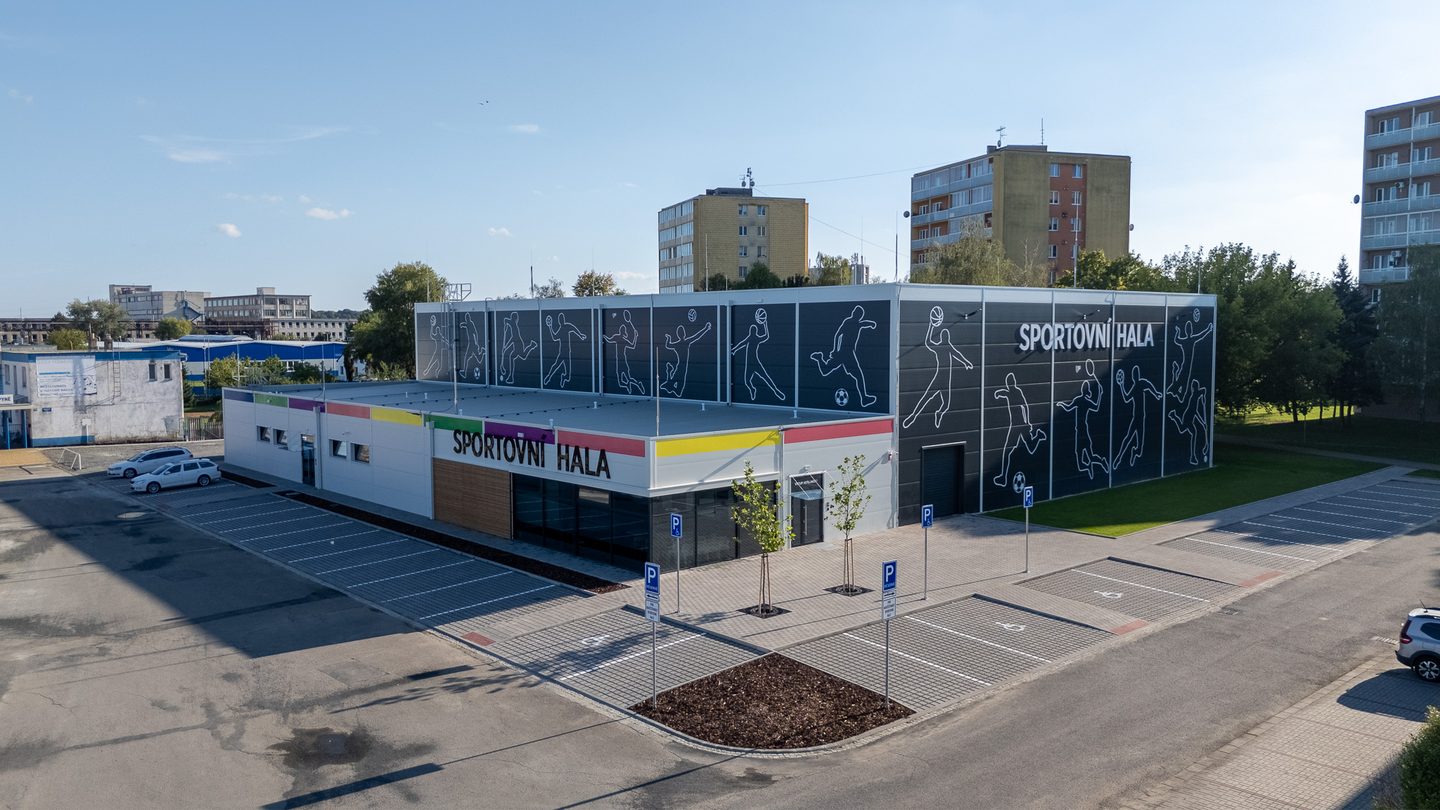 Sportovní hala z ptačí perspektivy: antracitová budova s bílými siluetami sportovců a nápisem SPORTOVNÍ HALA, před ní nové parkoviště.